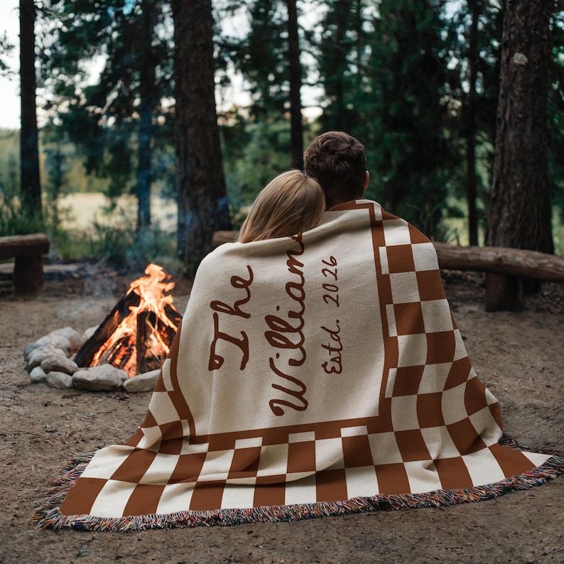 May include: A couple is wrapped in a cream and brown checkered blanket with the words "The Williams Estd. 2026" in a forest setting. The blanket has a fringed edge. A campfire is visible in the background.