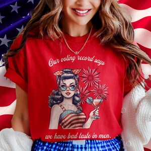 May include: Red t-shirt with a graphic of a woman in a patriotic outfit holding a cocktail, with text that reads "Our voting record says we have bad taste in men."