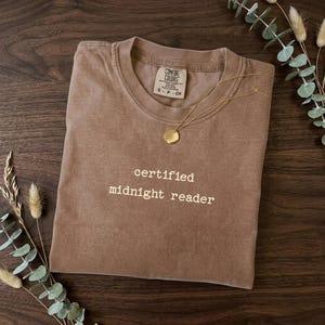 May include: A brown t-shirt with the words "certified midnight reader" printed in white. A gold necklace with a round pendant rests on the shirt. The shirt is folded on a wooden surface.