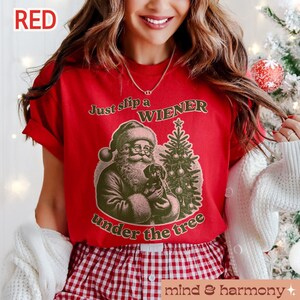 May include: Red t-shirt with a Christmas-themed graphic featuring Santa Claus holding a dachshund and a Christmas tree. The text on the shirt reads "Just slip a WIENER under the tree".