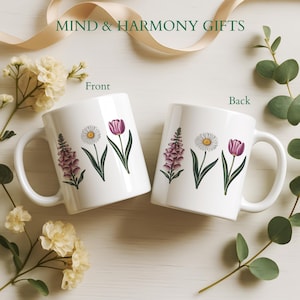 May include: Two white ceramic mugs with floral designs. One mug features a purple tulip, a daisy, and a purple flower. The other mug has the same floral arrangement. The text "MIND & HARMONY GIFTS" is at the top.