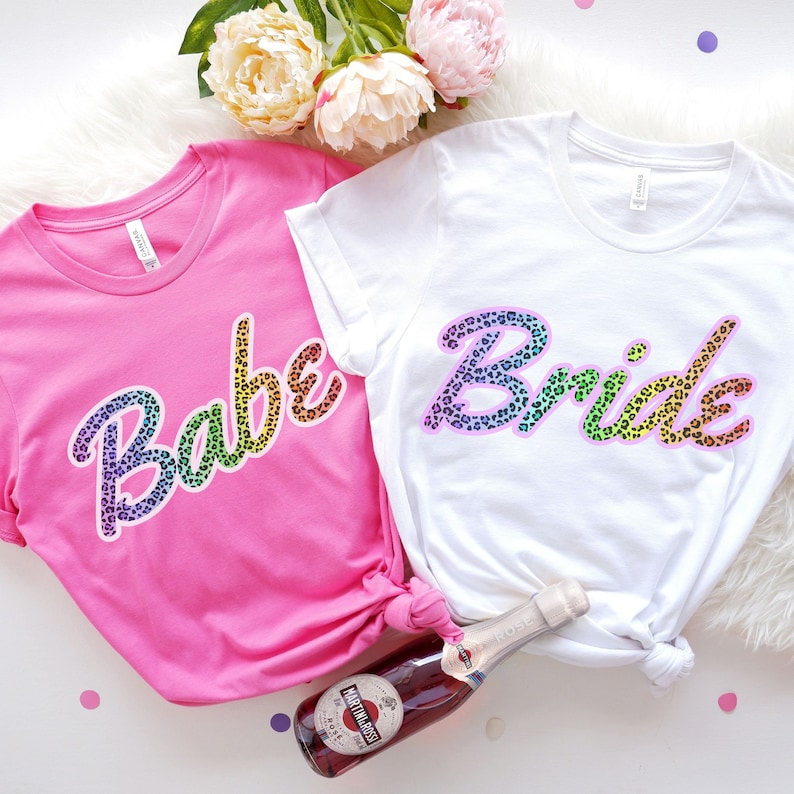 May include: Two white t-shirts with rainbow leopard print text. One shirt says "Babe" and the other says "Bride".