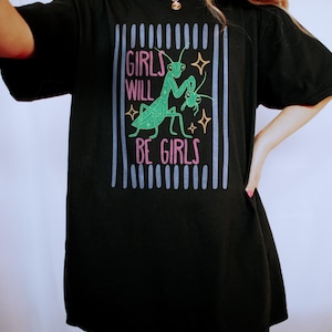 Puede incluir: Camiseta negra con el texto "GIRLS WILL BE GIRLS" en rosa y verde, junto con un gr&aacute;fico de una mantis religiosa. El dise&ntilde;o est&aacute; enmarcado por l&iacute;neas verticales azules y grises.