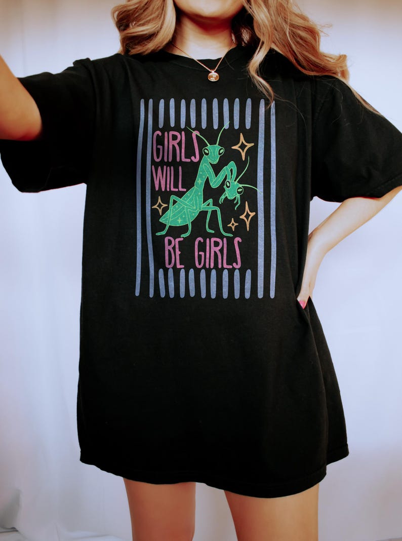 Puede incluir: Camiseta negra con el texto "GIRLS WILL BE GIRLS" en rosa y verde, junto con un gr&aacute;fico de una mantis religiosa. El dise&ntilde;o est&aacute; enmarcado por l&iacute;neas verticales azules y grises.