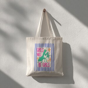 Puede incluir: Bolso tote de color crema con un gr&aacute;fico azul y rosa. El gr&aacute;fico presenta una ilustraci&oacute;n de mantis religiosa verde y el texto "GIRLS WILL BE GIRLS" en rosa y azul. La bolsa cuelga de un gancho de madera en una pared blanca.
