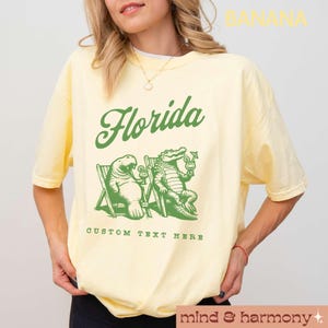 May include: A yellow t-shirt with a green graphic of a manatee and alligator sitting on beach chairs with drinks. The text "Florida" is above the graphic and "Custom Text Here" is below the graphic.
