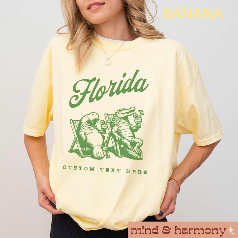 May include: A yellow t-shirt with a green graphic of a manatee and alligator sitting on beach chairs with drinks. The text "Florida" is above the graphic and "Custom Text Here" is below the graphic.