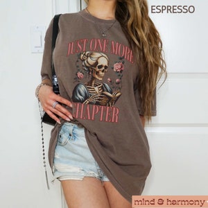 May include: A brown t-shirt with a graphic of a skeleton reading a book with the text "Just One More Chapter" printed on it.
