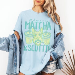 May include: Light blue t-shirt with the text "all I need is my MATCHA & SCOTTIE" in green. The design includes a cartoon image of a Scottish Terrier dog and two matcha drinks. The shirt is a casual, comfortable style.