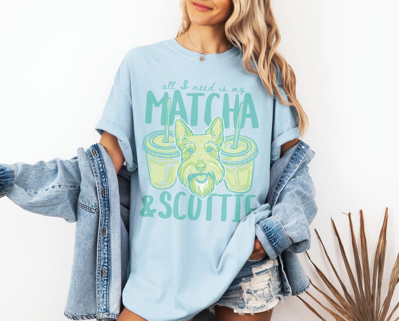May include: Light blue t-shirt with the text "all I need is my MATCHA & SCOTTIE" in green. The design includes a cartoon image of a Scottish Terrier dog and two matcha drinks. The shirt is a casual, comfortable style.