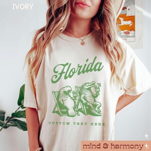 May include: Ivory t-shirt with a green graphic of a manatee and alligator sitting in beach chairs with drinks. The text "Florida" is above the graphic and "CUSTOM TEXT HERE" is below. The text "mind & harmony" is at the bottom of the image.