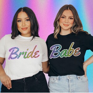 May include: Two white t-shirts with the words "Bride" and "Babe" in rainbow leopard print. The shirts are worn by two people with dark hair and are against a rainbow background.