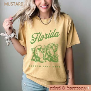 May include: A mustard yellow t-shirt with a green graphic of a manatee and alligator sitting in chairs with drinks. The text "Florida" is written in a green cursive font above the graphic. The text "CUSTOM TEXT HERE" is written below the graphic.