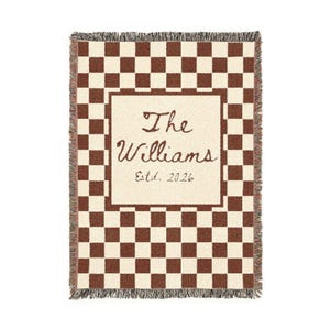 May include: A woven throw blanket with a brown and cream checkerboard pattern. The center features a cream-colored rectangle with the text "The Williams Estd. 2026" in brown script. The edges have a fringed trim.