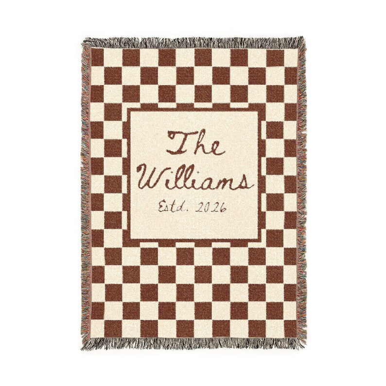 May include: A woven throw blanket with a brown and cream checkerboard pattern. The center features a cream-colored rectangle with the text "The Williams Estd. 2026" in brown script. The edges have a fringed trim.
