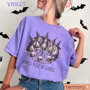 Peut inclure: T-shirt violet avec un motif de quatre sorci&egrave;res assises autour d'une table avec des cr&acirc;nes et des os. Le texte "Girls Will Be Girls" est imprim&eacute; sous le motif.