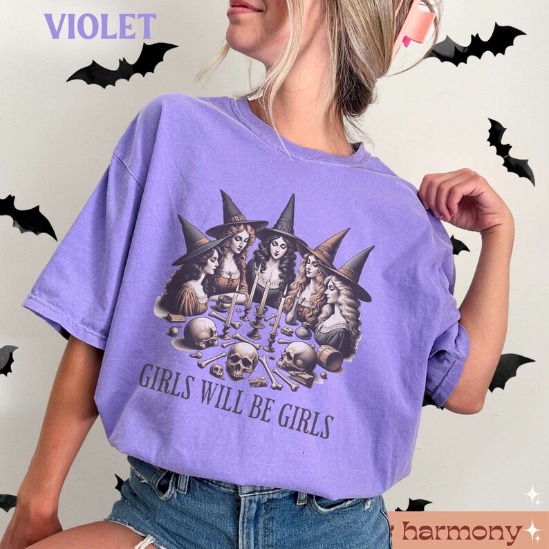 Peut inclure: T-shirt violet avec un motif de quatre sorci&egrave;res assises autour d'une table avec des cr&acirc;nes et des os. Le texte "Girls Will Be Girls" est imprim&eacute; sous le motif.