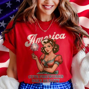 May include: Red t-shirt with the word "America" in white script and a vintage illustration of a woman holding a sparkler. The text "CUTE FIREWORKS UGLY REALITY" is below the illustration.