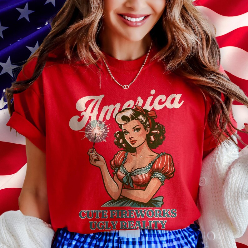 May include: Red t-shirt with the word "America" in white script and a vintage illustration of a woman holding a sparkler. The text "CUTE FIREWORKS UGLY REALITY" is below the illustration.