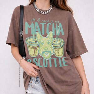May include: Brown t-shirt with the text "all I need is my MATCHA & SCOTTIE" in teal and a graphic of a dog and two matcha drinks. The shirt has short sleeves and a relaxed fit.