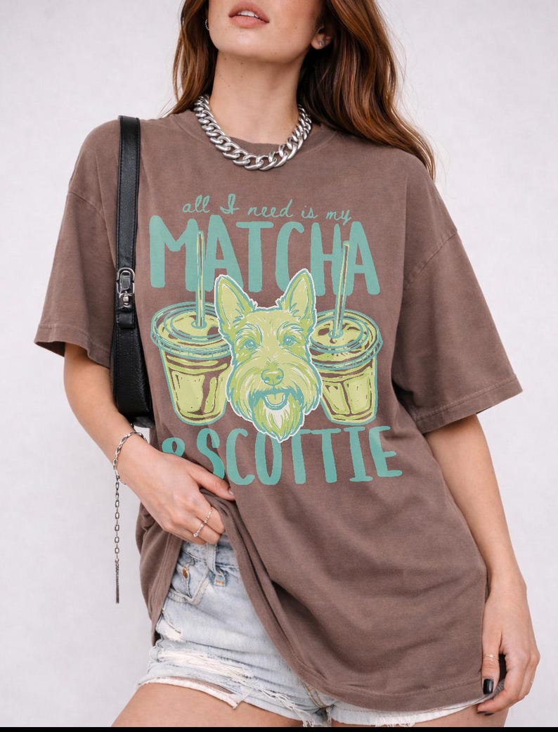 May include: Brown t-shirt with the text "all I need is my MATCHA & SCOTTIE" in teal and a graphic of a dog and two matcha drinks. The shirt has short sleeves and a relaxed fit.