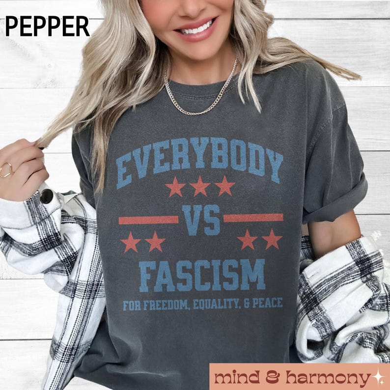 Puede incluir: Una camiseta gris oscuro con un gr&aacute;fico de estilo vintage que dice "Everybody vs Fascism" con estrellas rojas y rayas rojas y blancas. El texto debajo del gr&aacute;fico dice "For Freedom, Equality, & Peace".