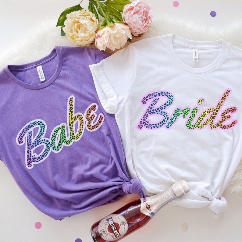 May include: Two t-shirts with a rainbow leopard print design. One shirt says "Babe" and the other says "Bride".