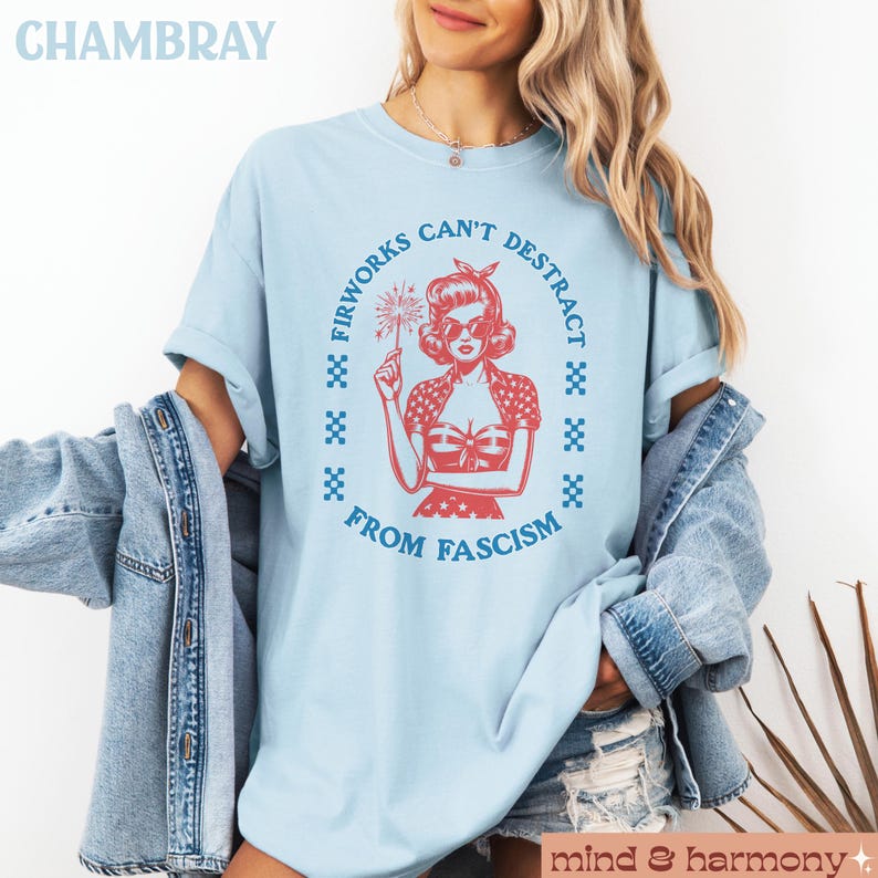 May include: Pale blue t-shirt featuring a red graphic of a woman with a firework, encircled by the words "Fireworks Can't Distract From Fascism." The word "CHAMBRAY" is at the top left.