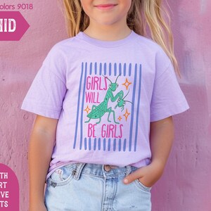 Puede incluir: Una camiseta lila de manga corta para j&oacute;venes con el texto "GIRLS WILL BE GIRLS" y un dise&ntilde;o de mantis religiosa verde. La camiseta est&aacute; etiquetada como "ORCHID" y forma parte de la colecci&oacute;n "MIND & HARMONY GIFTS".