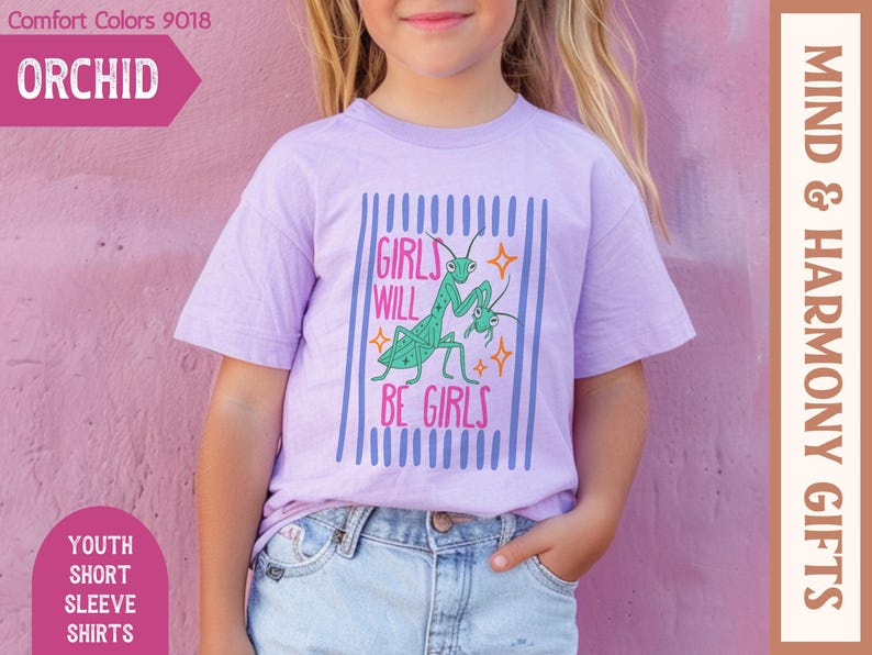 Puede incluir: Una camiseta lila de manga corta para j&oacute;venes con el texto "GIRLS WILL BE GIRLS" y un dise&ntilde;o de mantis religiosa verde. La camiseta est&aacute; etiquetada como "ORCHID" y forma parte de la colecci&oacute;n "MIND & HARMONY GIFTS".