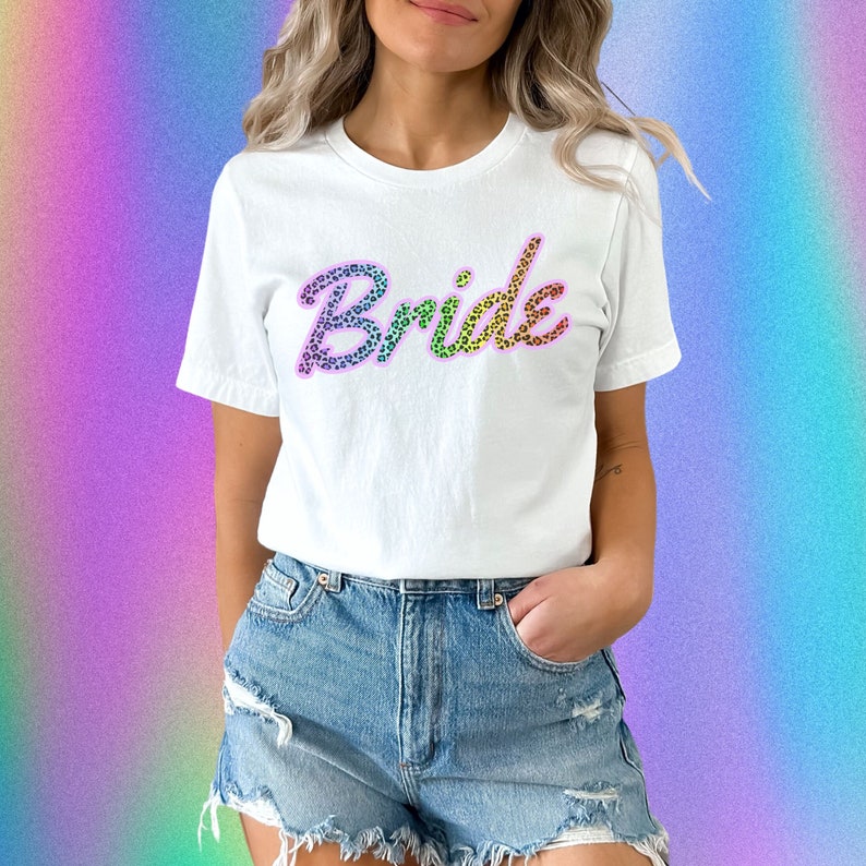 May include: White t-shirt with the word "bride" written in rainbow colors with a leopard print pattern.