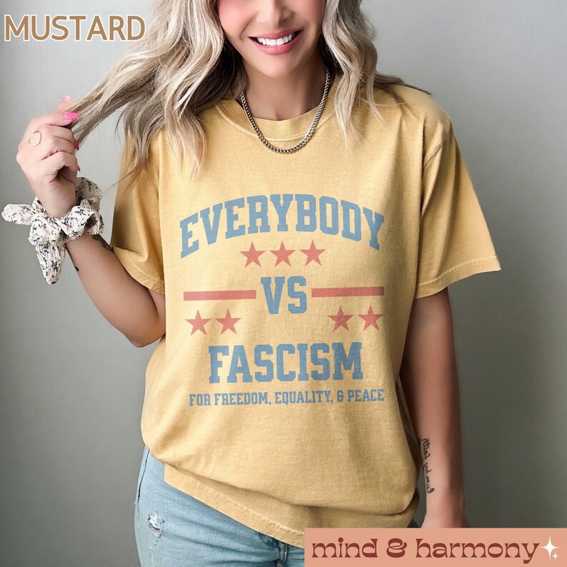 Puede incluir: Una camiseta amarilla mostaza con el texto "EVERYBODY VS FASCISM" en azul y rojo. El texto est&aacute; en una fuente de estilo vintage e incluye estrellas y rayas. El texto debajo del texto principal dice "FOR FREEDOM, EQUALITY, & PEACE".