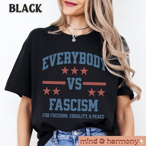 Puede incluir: Camiseta negra con el texto "EVERYBODY VS FASCISM" en letras azules y rojas. El texto est&aacute; rodeado de estrellas y una franja roja. El texto "FOR FREEDOM, EQUALITY, & PEACE" est&aacute; impreso debajo del texto principal.