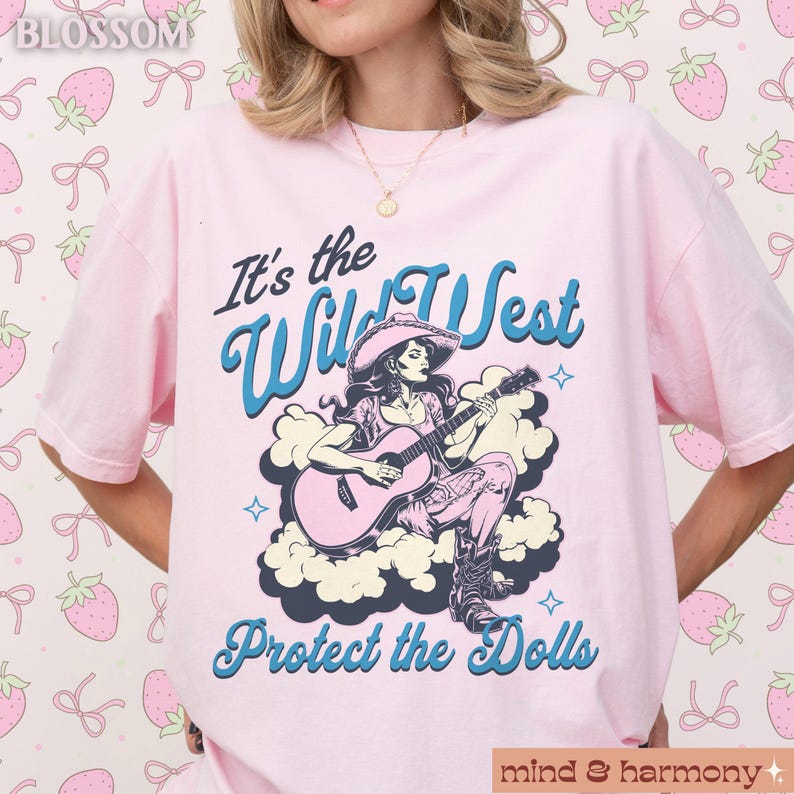Może przedstawiać: R&oacute;żowy T-shirt z grafiką kobiety grającej na gitarze z tekstem "It's the Wild West Protect the Dolls" na r&oacute;żowym tle.