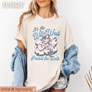 Może przedstawiać: Koszulka w kolorze kości słoniowej z grafiką kobiety grającej na gitarze z tekstem "It's the Wild West Protect the Dolls".