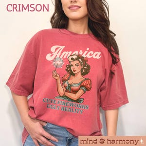 May include: Crimson-colored t-shirt with the word "America" above a vintage illustration of a woman holding a sparkler. The text "CUTE FIREWORKS UGLY REALITY" is below the illustration. The shirt is part of the "mind & harmony" collection.