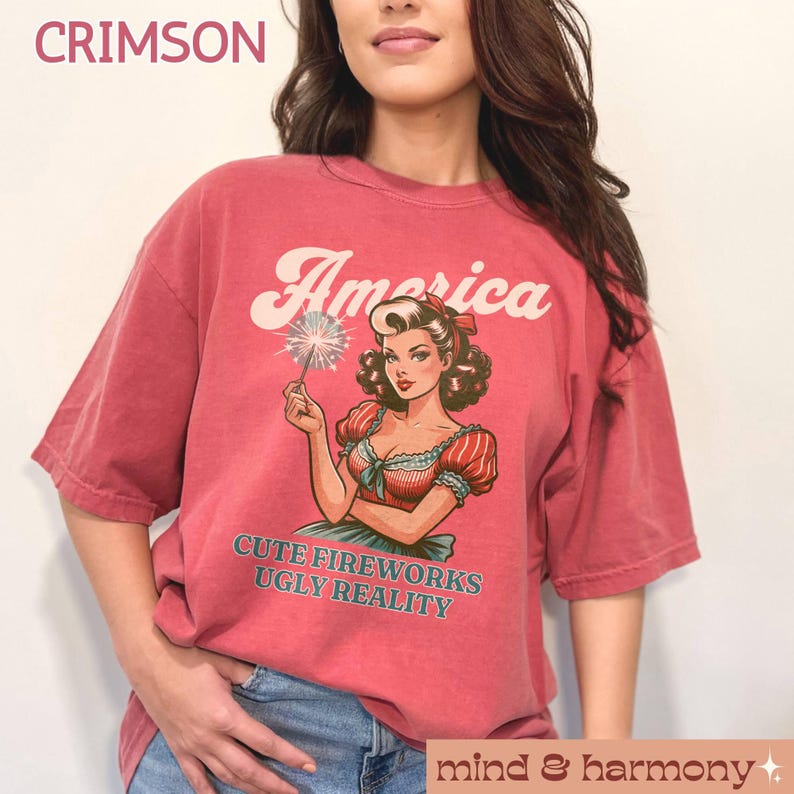 May include: Crimson-colored t-shirt with the word "America" above a vintage illustration of a woman holding a sparkler. The text "CUTE FIREWORKS UGLY REALITY" is below the illustration. The shirt is part of the "mind & harmony" collection.