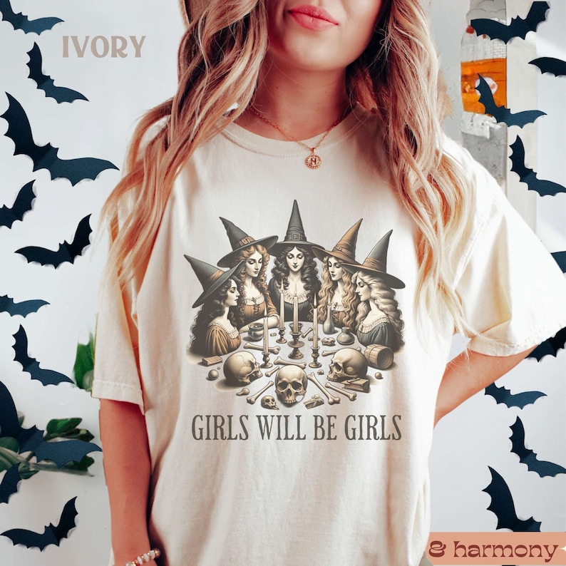 Peut inclure: Un t-shirt blanc avec un motif de quatre sorci&egrave;res portant des chapeaux et des robes noirs, r&eacute;unies autour d'une table avec des cr&acirc;nes et des os. Le texte "Girls Will Be Girls" est imprim&eacute; sous le motif.