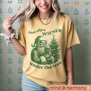 May include: A mustard yellow t-shirt with a green graphic of Santa Claus holding a dachshund under a Christmas tree. The text "Just slip a WIENER under the tree" is printed on the shirt.