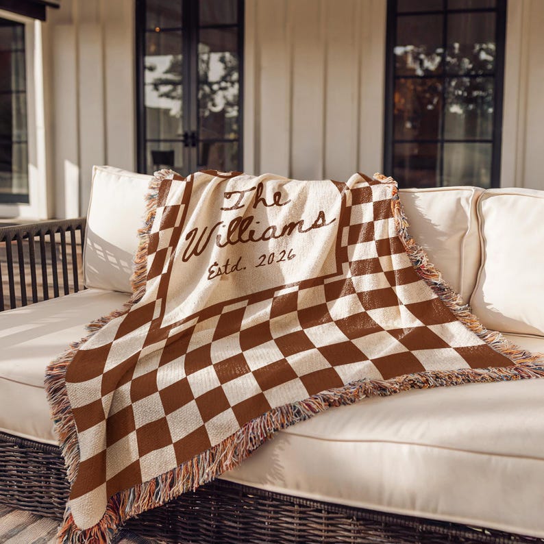 May include: A woven throw blanket with a brown and cream checkerboard pattern. The blanket is draped over a white sofa and features the text "The Williams Estd. 2026" in a script font. The edges have a fringed trim.