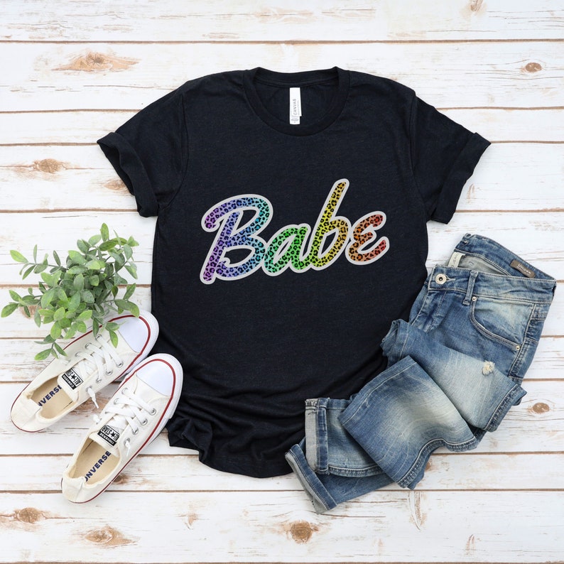 May include: A black t-shirt with the word "Babe" written in rainbow colors with a leopard print pattern. The word is in a cursive font.