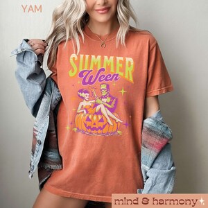 May include: Orange t-shirt with the text "Summerween" in green and purple. The graphic features the Bride of Frankenstein and Frankenstein sitting on a jack-o'-lantern, holding cocktails. The shirt has a vintage, distressed look.