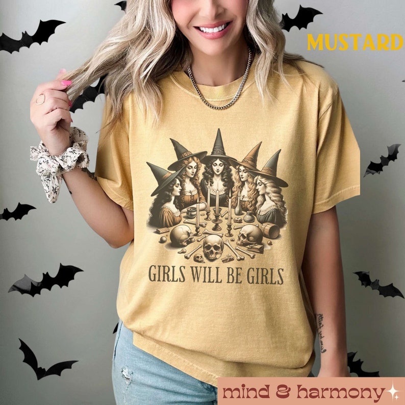 Peut inclure: Un t-shirt jaune moutarde avec une illustration vintage de quatre sorci&egrave;res r&eacute;unies autour d'une table avec des bougies et des cr&acirc;nes. Le texte "Girls Will Be Girls" est imprim&eacute; sous l'illustration.
