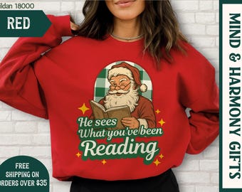 May include: A red sweatshirt featuring a cartoon Santa Claus reading a book, with the text "He sees What you've been Reading". The Gildan 18000 sweatshirt has green and yellow accents. A festive design for the Christmas season.