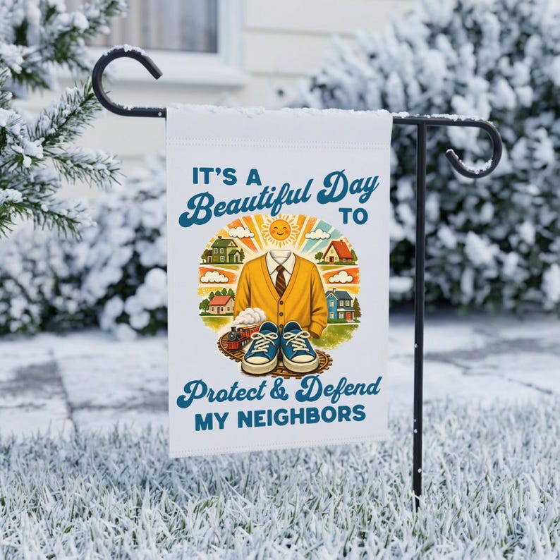 May include: A white garden flag with the text "It's a Beautiful Day to Protect & Defend MY NEIGHBORS." The flag features a cartoon illustration of a yellow sweater, houses, a train, and blue sneakers. The flag is displayed on a black metal stand.