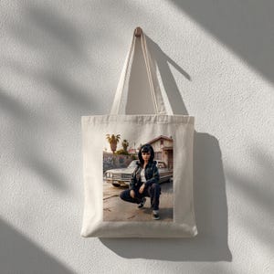 May include: A cream-colored tote bag with a printed image of a person squatting in front of a classic car. The person is wearing a jacket, pants, and sneakers. The background includes a building and palm trees.