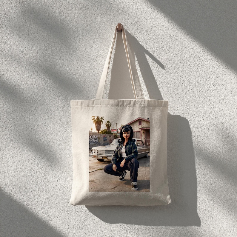 May include: A cream-colored tote bag with a printed image of a person squatting in front of a classic car. The person is wearing a jacket, pants, and sneakers. The background includes a building and palm trees.