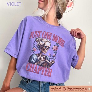 May include: A purple t-shirt with a graphic of a skeleton reading a book. The text on the shirt reads "Just One More Chapter".