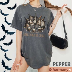 Peut inclure: Un t-shirt gris fonc&eacute; avec un imprim&eacute; graphique de cinq sorci&egrave;res portant des chapeaux et des robes noirs, tenant des bougies et des cr&acirc;nes. Le texte "Girls Will Be Girls" est imprim&eacute; sous le graphique.
