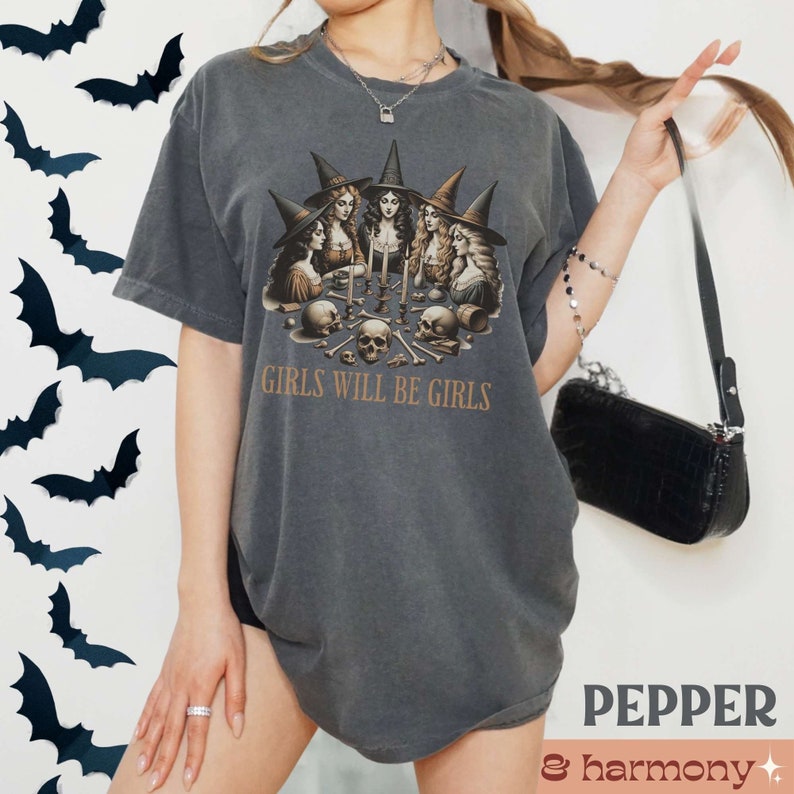 Peut inclure: Un t-shirt gris fonc&eacute; avec un imprim&eacute; graphique de cinq sorci&egrave;res portant des chapeaux et des robes noirs, tenant des bougies et des cr&acirc;nes. Le texte "Girls Will Be Girls" est imprim&eacute; sous le graphique.